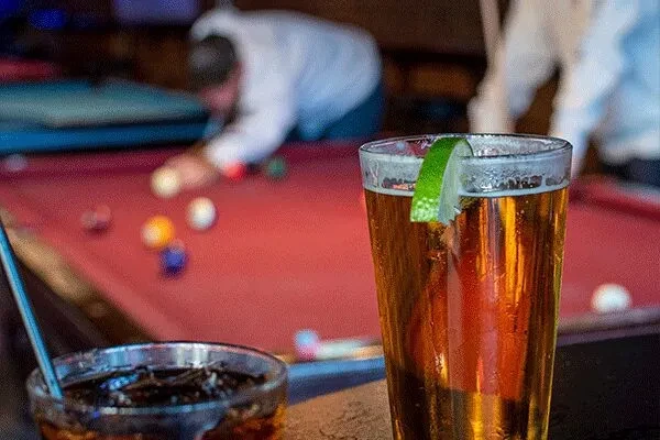 Amber beer with lime, dark drink, pool game in background.