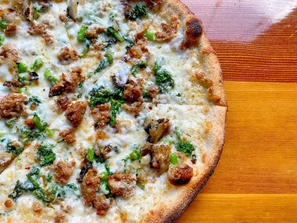 Sliced pizza with browned sausage, green vegetables, melted cheese on wood.