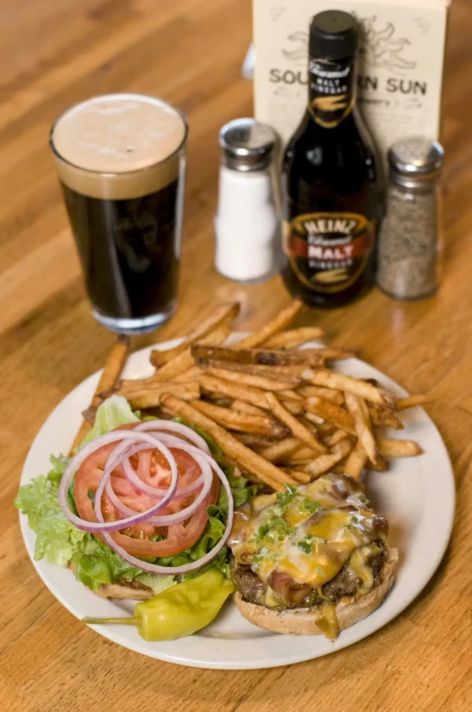 Overhead view: open-faced burger, fries, dark beer on wooden table.