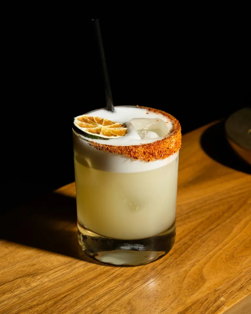 Pale green cocktail with foam, dried citrus, and chili-spiced rim.
