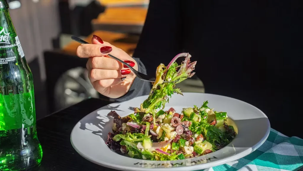 Hand with red nails eats a salad of greens, crumbled cheese, and olives.