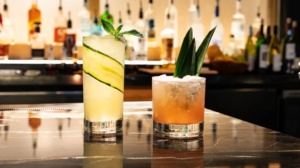 Two garnished cocktails: yellow with cucumber, amber with foam, on bar.