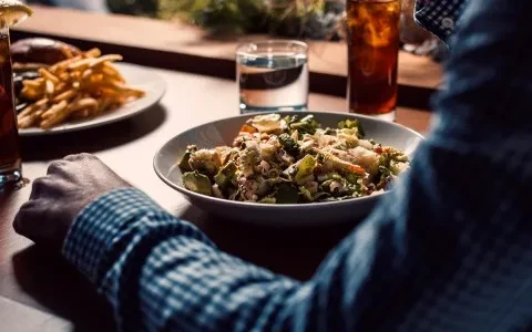 Colorful salad bowl, fries, drinks on table, viewed over person's shoulder.