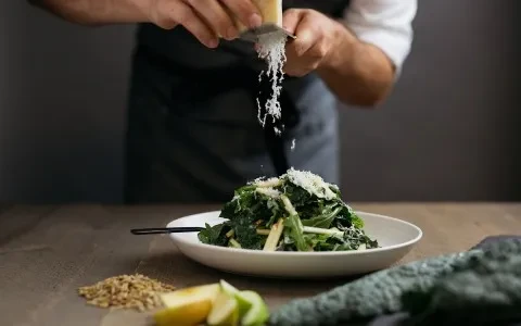 Person's hands grating cheese onto fresh green salad in white bowl.