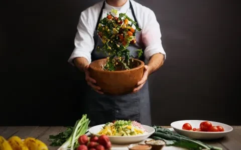Chef tosses vibrant salad in wooden bowl; fresh ingredients on table.