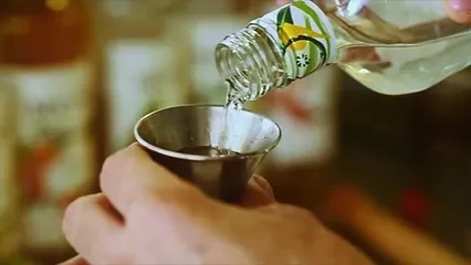 Hands pour clear liquid from a green-labeled bottle into a silver jigger.