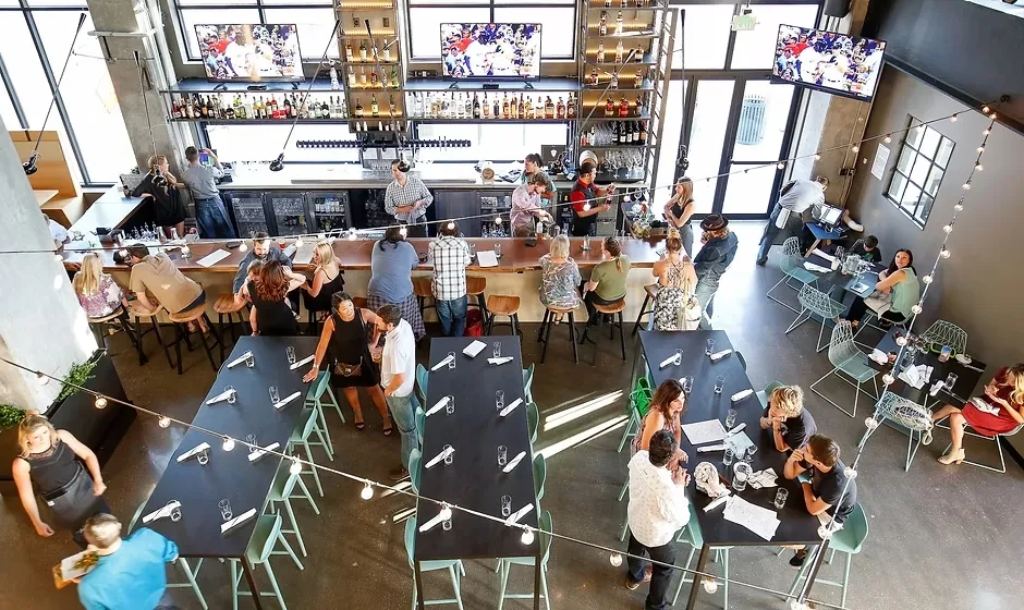 Bustling industrial-chic restaurant interior with bar, diners, and sports on TVs.