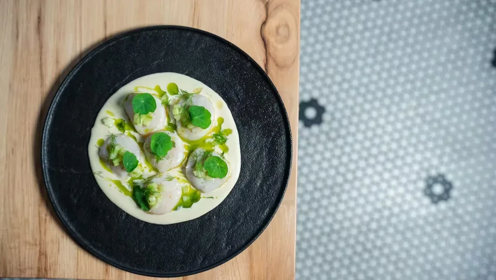 Five scallops in creamy sauce with green garnish on dark plate.
