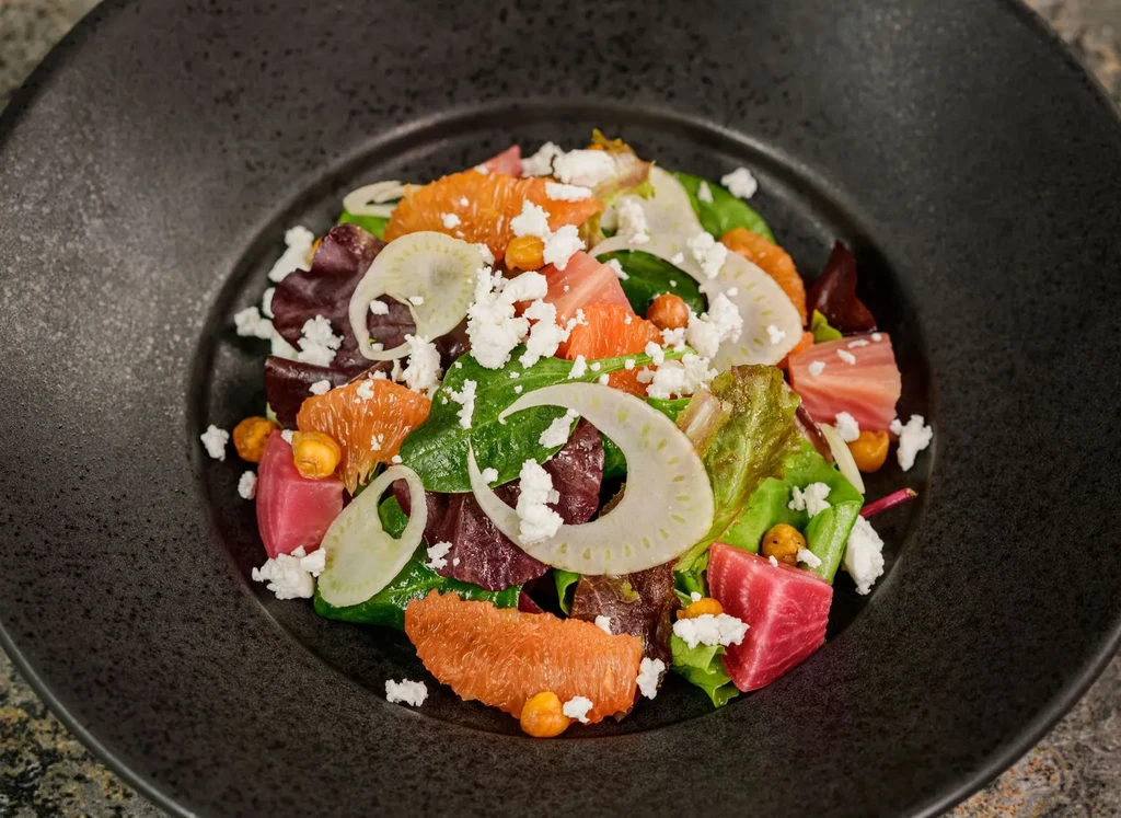 Colorful salad with greens, citrus, beets, and cheese in dark bowl.