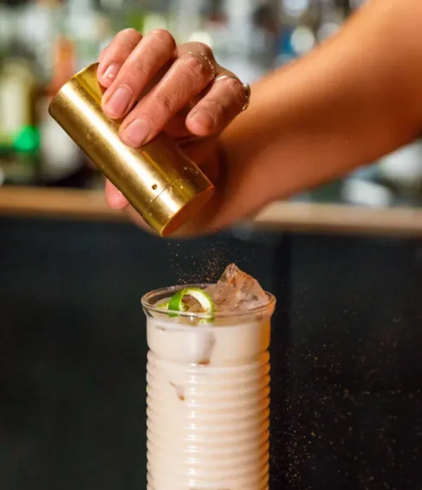 Hand sprinkles dark powder from golden shaker onto creamy cocktail.