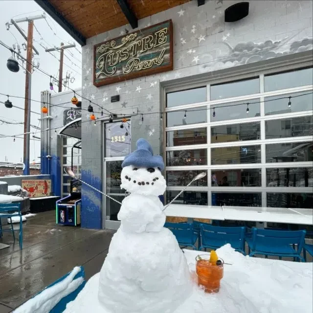 Snowman in blue beanie next to orange cocktail on snowy Lustre Pearl patio.