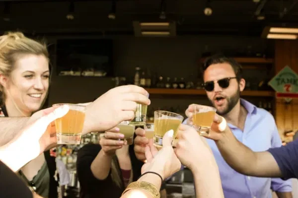 Hands clinking glasses in a toast, smiling woman, bearded man at bar.