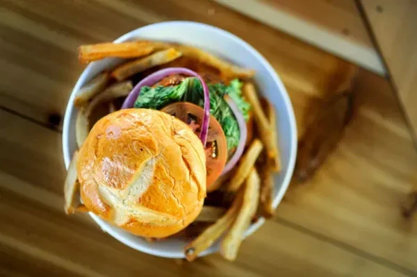 Top-down view: Hamburger with vegetables, fries in white bowl on wood.