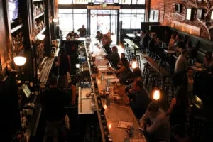Busy restaurant bar interior with patrons, staff, dark wood, warm lighting.