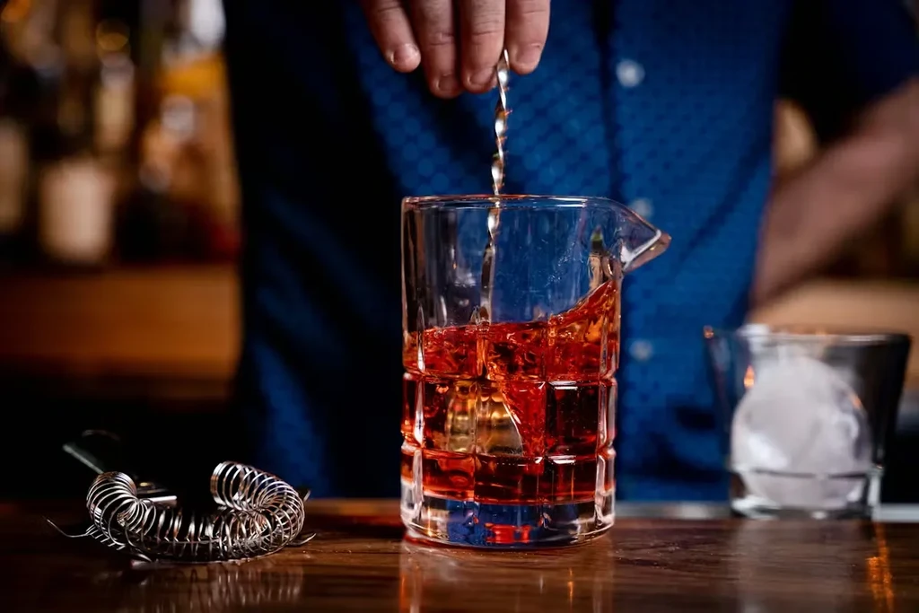 Bartender's hands stir reddish-orange drink with a spiral spoon in mixing glass.