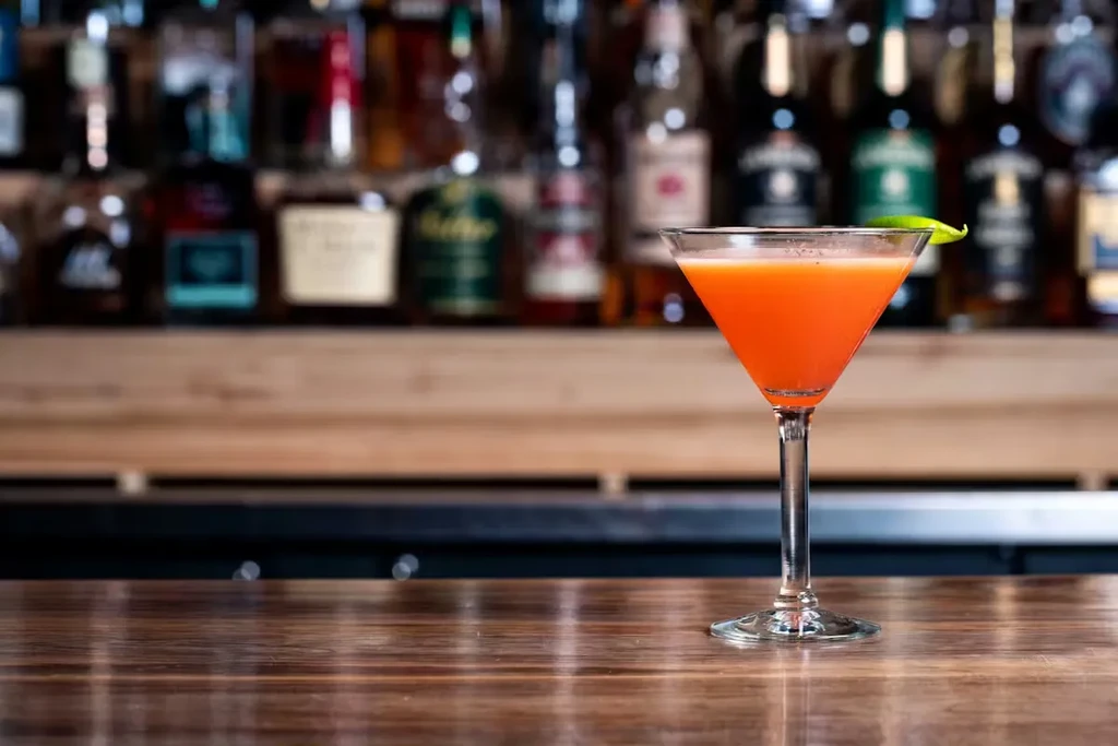 Orange-red cocktail in martini glass with green peel on a bar.
