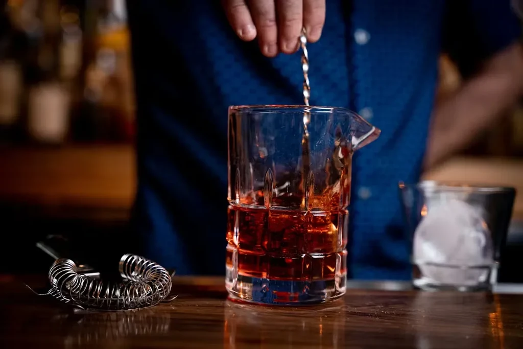 Bartender stirs a reddish-brown drink in a faceted mixing glass with ice.