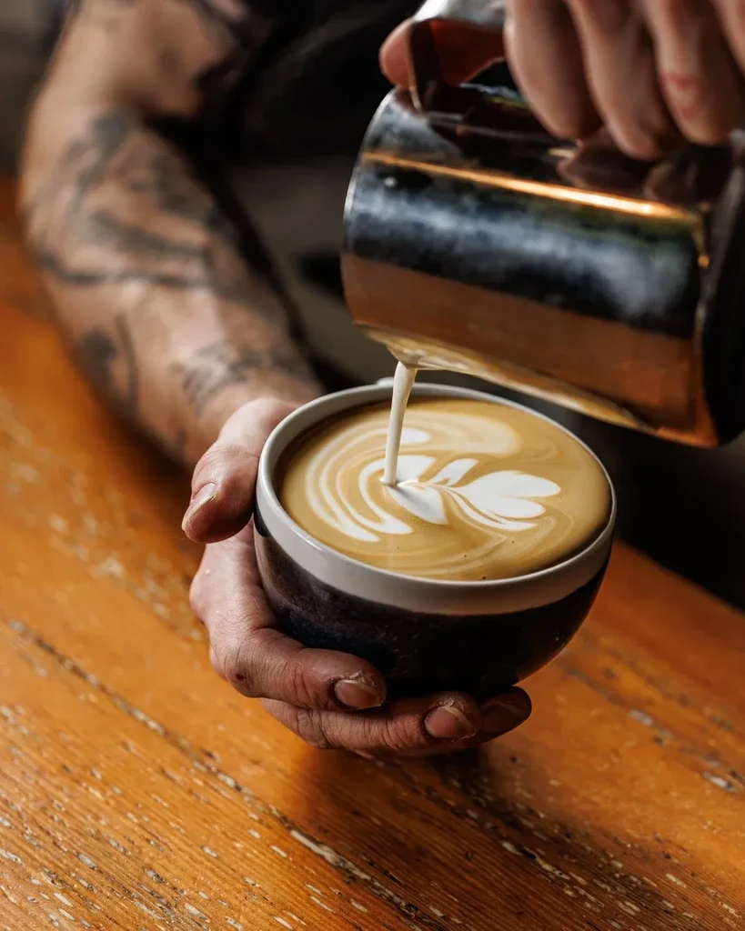 Barista's tattooed hands pour milk, creating rosetta latte art.