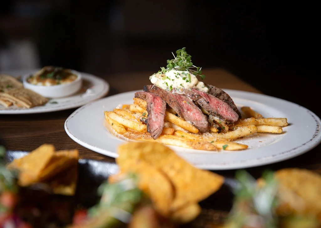 Close-up: Sliced medium-rare steak, fries, sauce, and microgreens on wood.