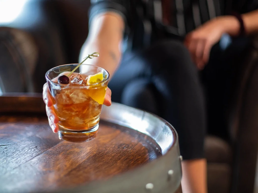 Amber cocktail with ice, cherry, lemon peel, held by hand.