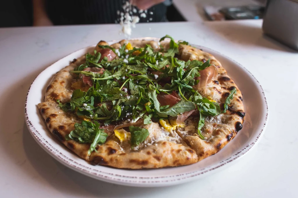 Hand sprinkles cheese over gourmet pizza with arugula and prosciutto.