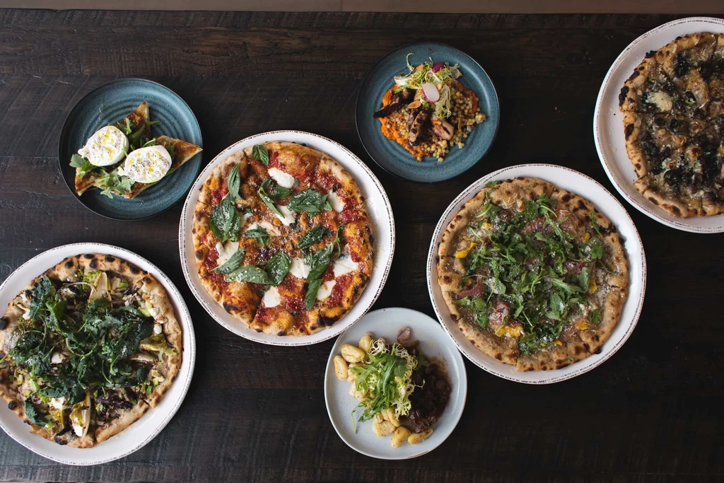Dark wooden table laden with various pizzas, burrata, octopus, and gnocchi.