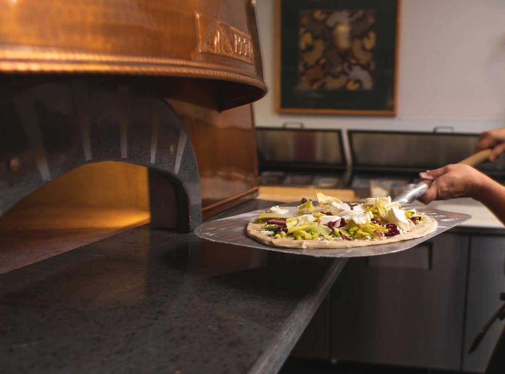 Hands insert uncooked pizza into a glowing, copper-colored oven.