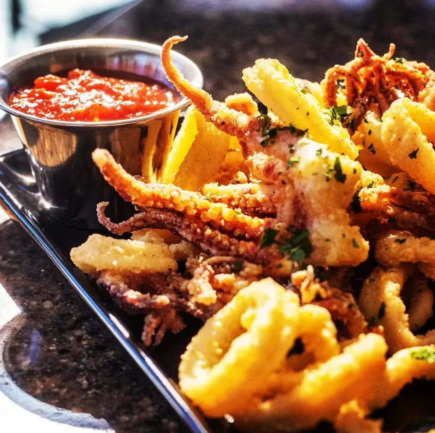 Fried calamari with herbs, red dipping sauce, and lemon on a black plate.