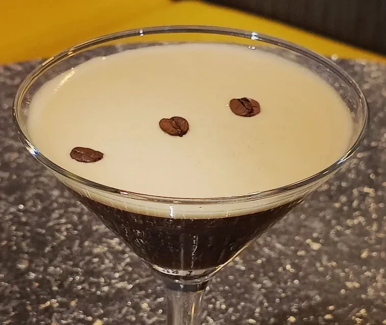 Layered dark drink in martini glass with foam and coffee beans.