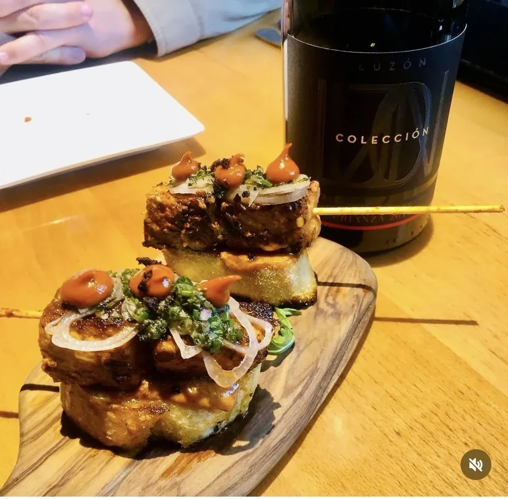 Grilled pintxos with herbs, sauce, on board next to red wine.