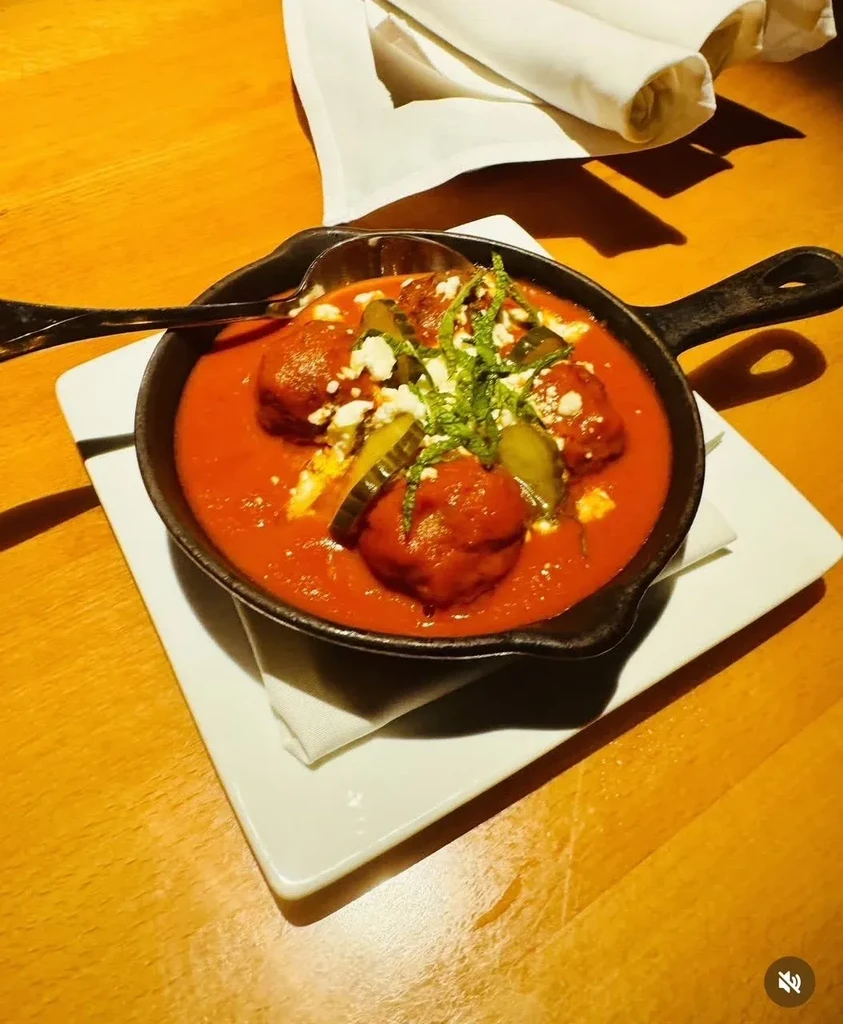 Skillet of meatballs in tomato sauce, garnished with cheese, pickles, herbs.