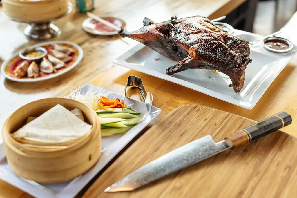 Roasted Peking duck, pancakes, vegetables, and dumplings on a wooden table.