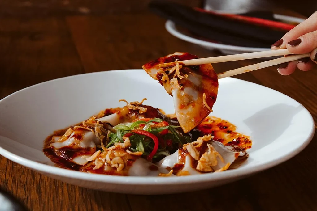 Chopsticks lift a saucy dumpling topped with green onions and chili.