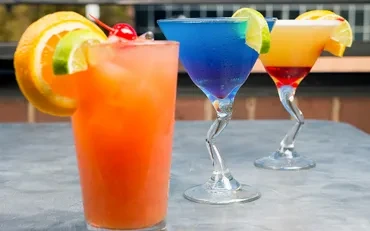 Three vibrant, garnished cocktails on grey counter, blurred background.