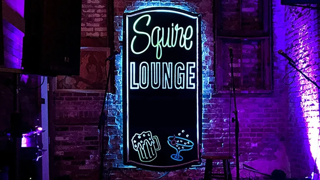 Vertical "Squire LOUNGE" neon sign on a purple-lit brick wall.