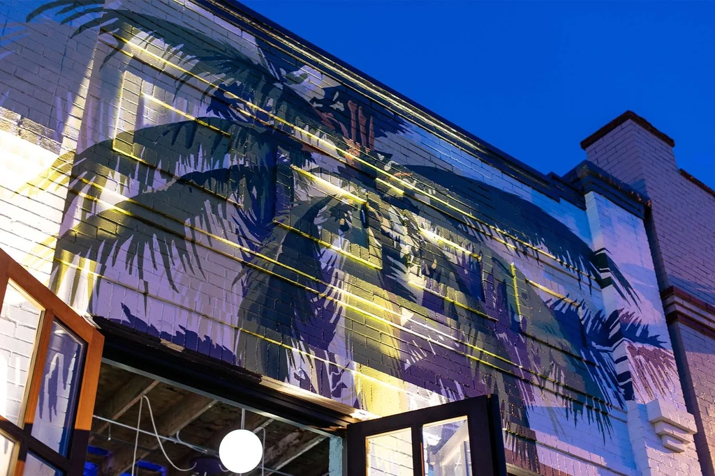 Dramatically lit palm frond mural on a white brick building at dusk.
