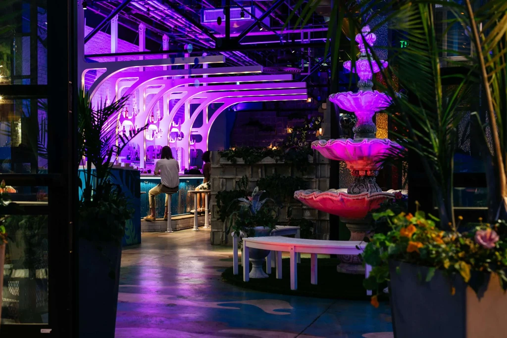 Purple-lit modern bar interior with wave design, fountain, and people.