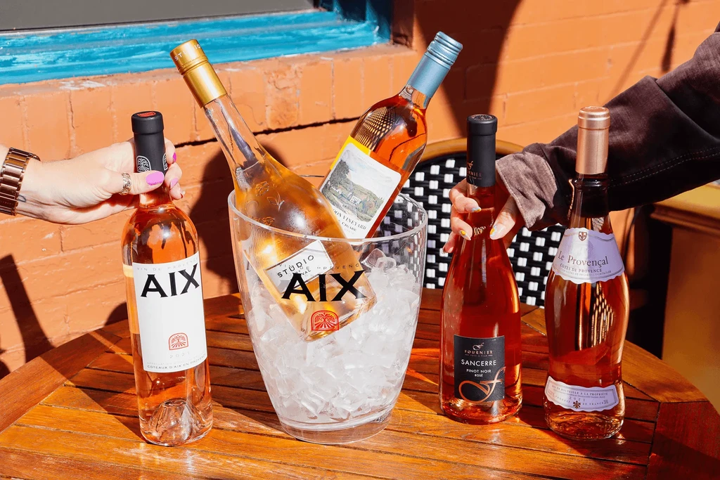 Hands holding rosé bottles on a sunny wooden table with ice.