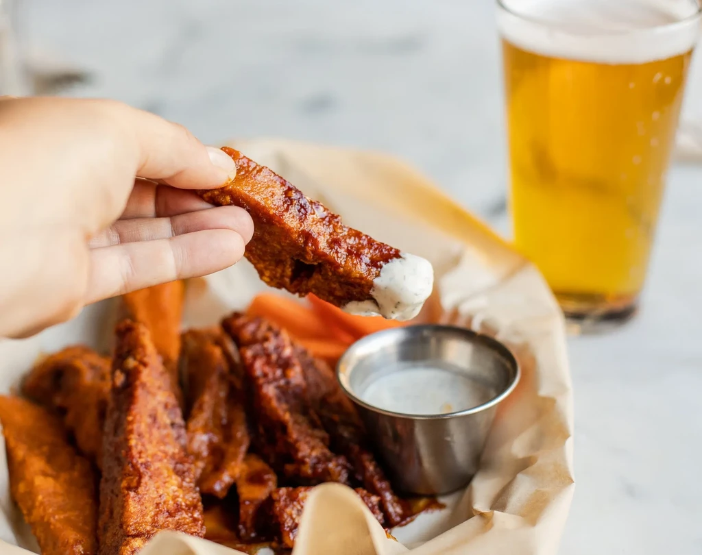 Hand holding a boneless wing dipped in creamy sauce with beer.