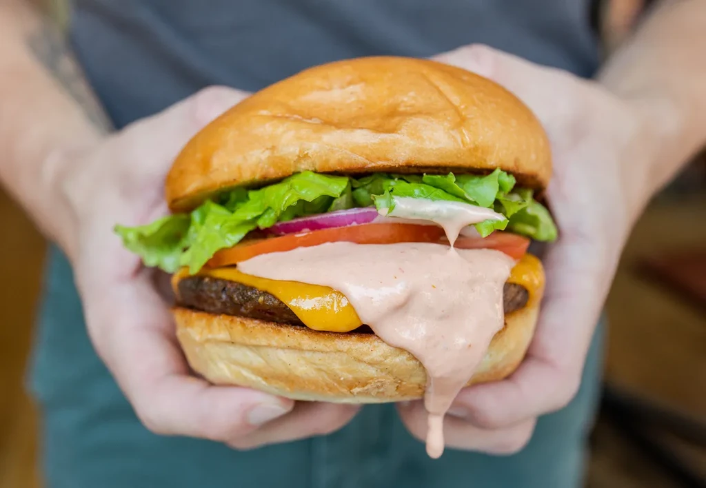 Hands hold a burger with melted cheese and dripping pink sauce.