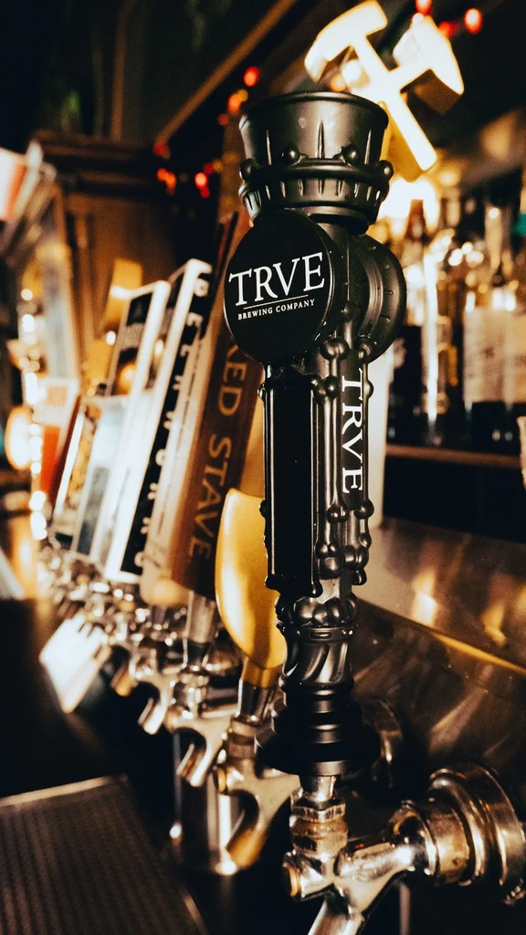 Ornate TRVE Brewing Company beer tap among other blurred bar taps.