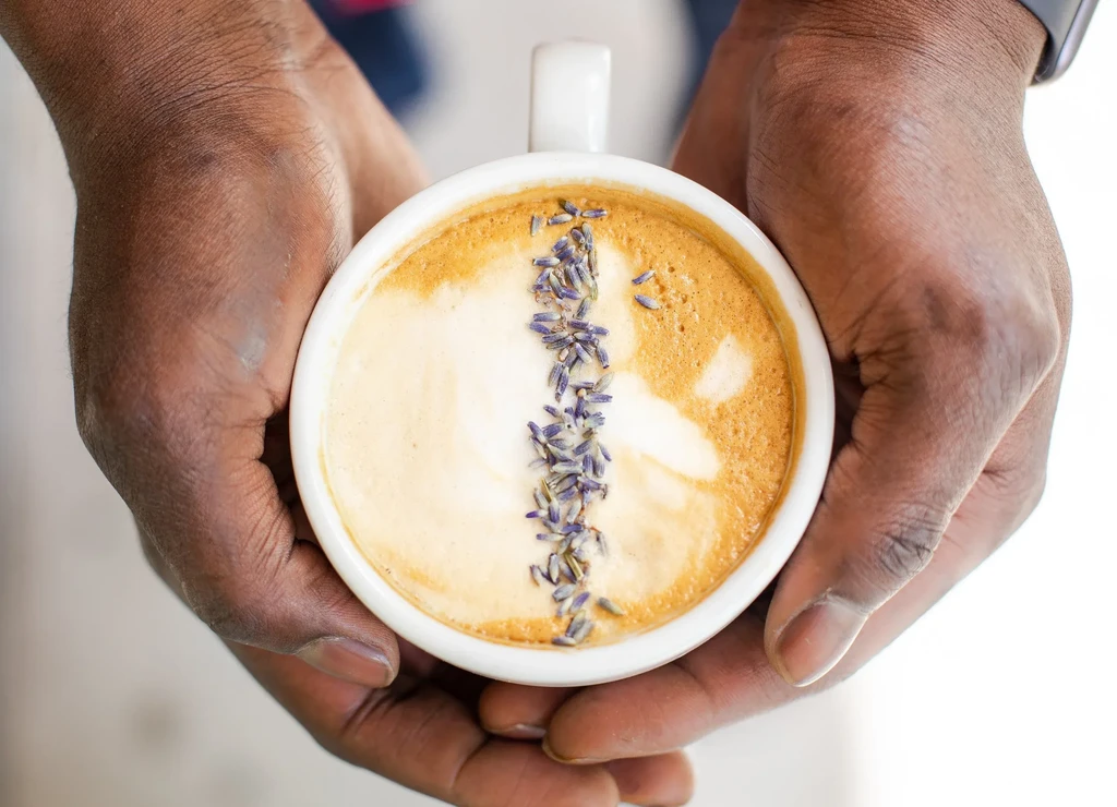 Top-down view of dark hands holding a lavender-garnished latte.