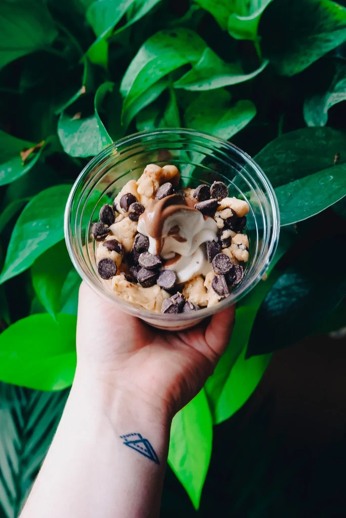 Hand holds clear cup of cookie dough, soft-serve, and chocolate.