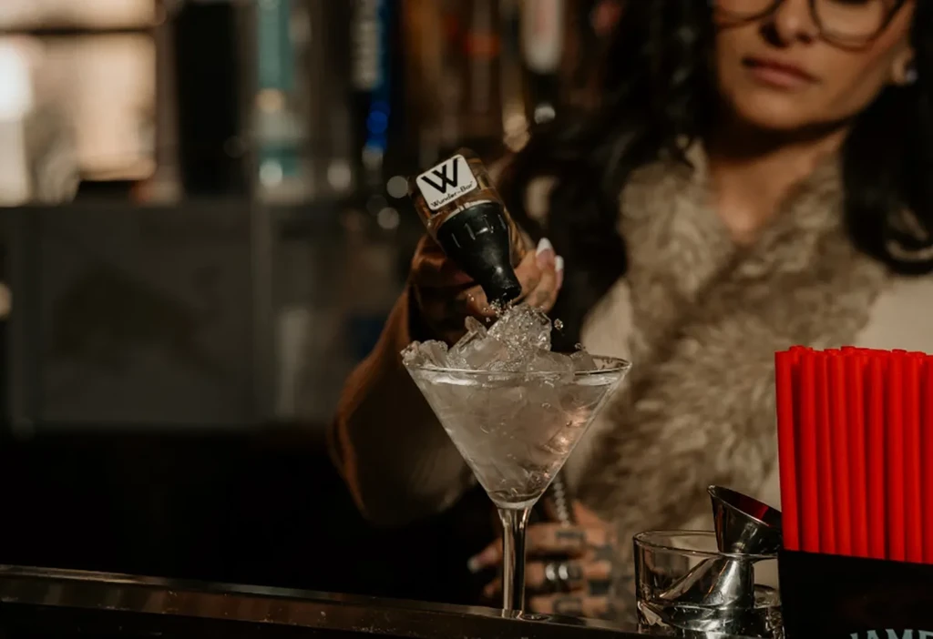 Person pours liquid from bottle into martini glass with splashing ice.
