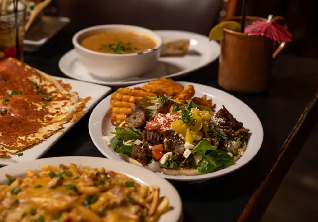 Table in a restaurant with cheesy fries, salad, soup, and cocktail.