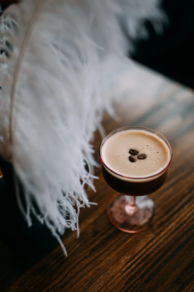Espresso martini in coupé glass with coffee beans, dark wood, feather.