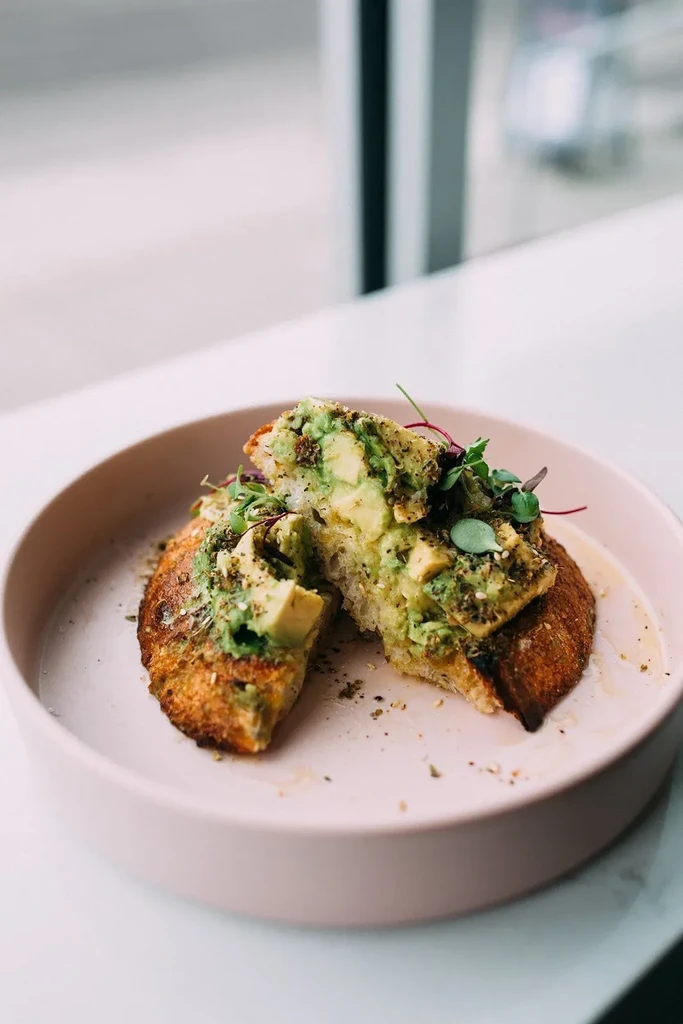 Rustic avocado toast on a pink plate with herbs and sprouts.