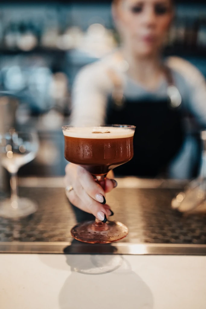 Hand holding dark frothy cocktail with garnish in a modern bar.