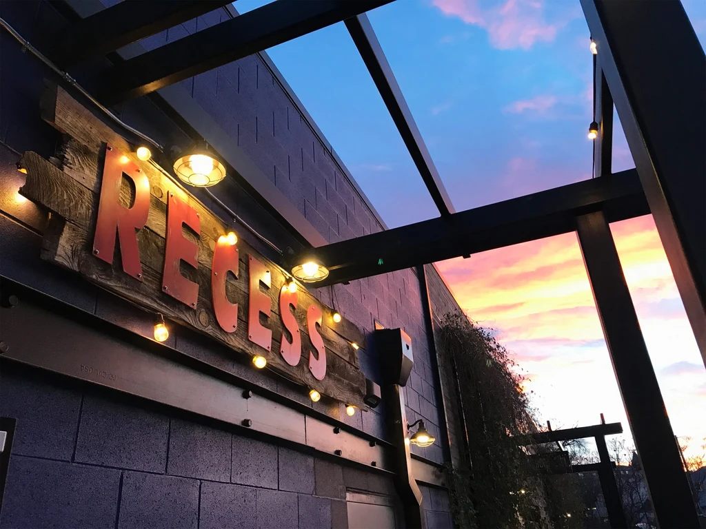 Rustic 'RECESS' sign illuminated by lights at sunset.