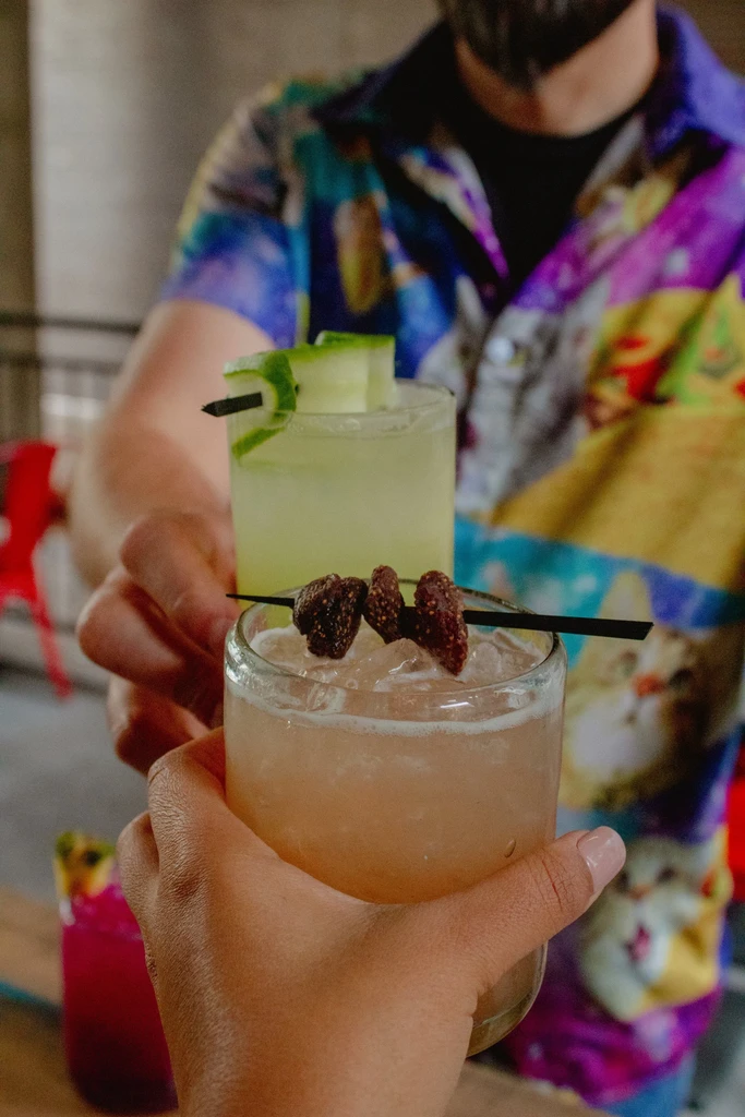 Two hands raise garnished peach and pale green cocktails in a toast.